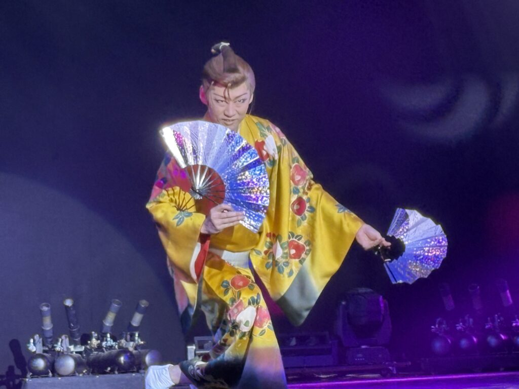 A male performer in a golden kimono decorated with floral patterns holding two fans during a dance number.