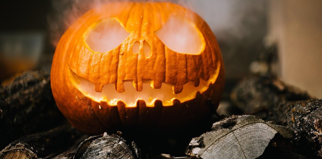 Carved jack-o’-lantern glowing with smoke coming from its mouth and eyes, placed on logs.