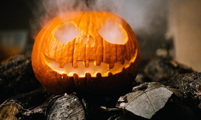 Carved jack-o’-lantern glowing with smoke coming from its mouth and eyes, placed on logs.