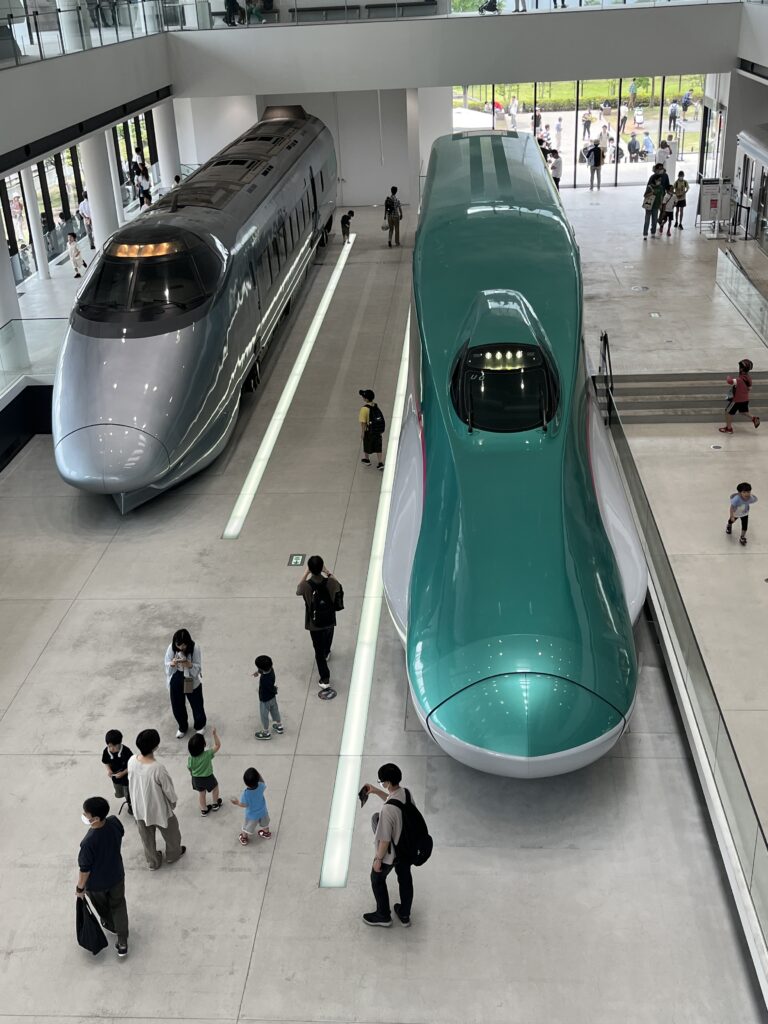 Two full-size Shinkansen train models displayed side by side in the Railway Museum’s main entrance hall, with visitors walking around the sleek silver and green bullet trains.