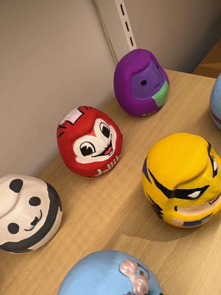 Hand-painted daruma dolls displayed on a wooden table, including a red daruma painted like Jollibee, a purple daruma with a simple face mask design, a yellow daruma with sharp anime-style eyes, a panda-themed daruma, and a blue daruma with a pink bow.