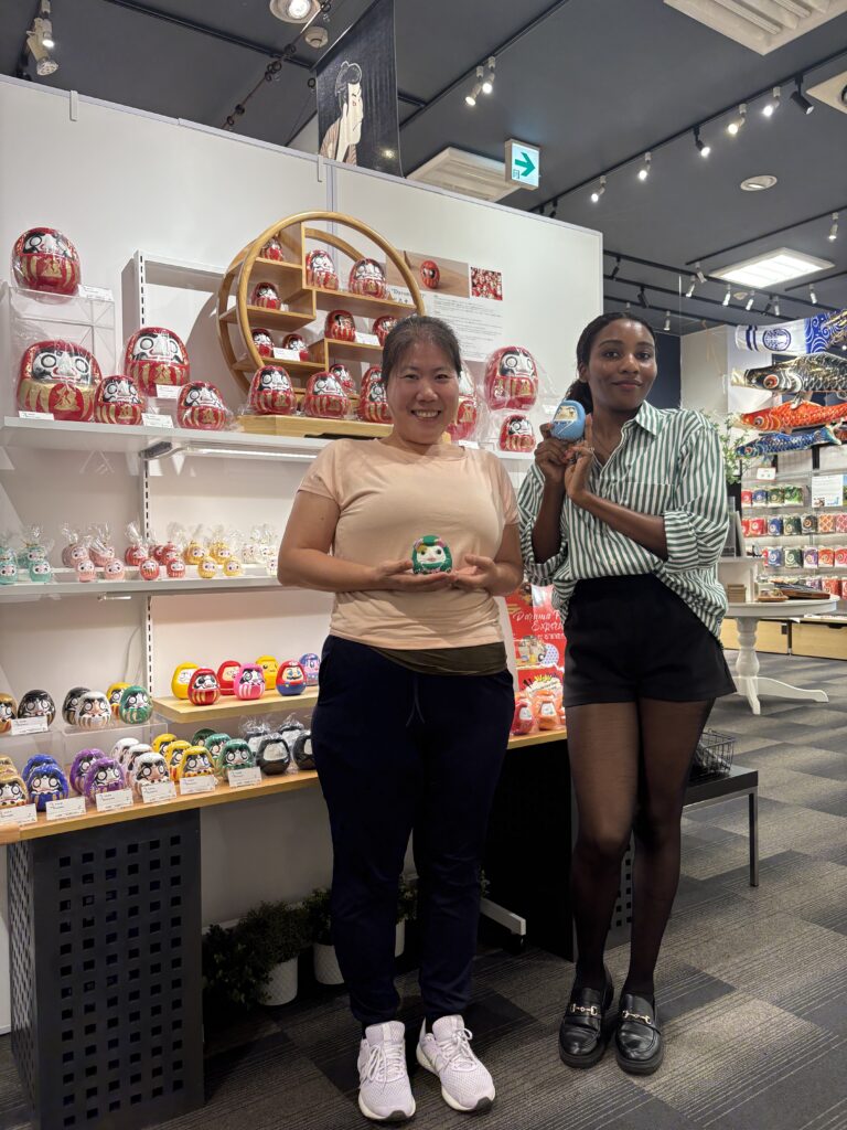 Two people standing inside a daruma shop, each holding a painted daruma doll, with shelves full of colorful daruma behind them.