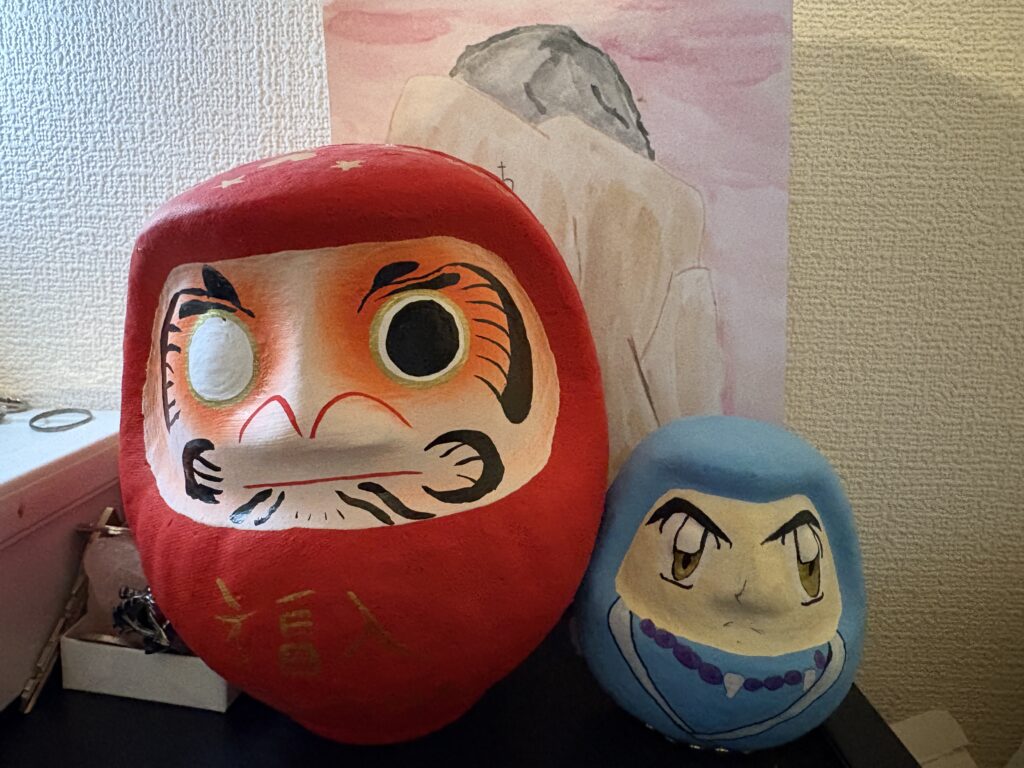 Two painted daruma dolls placed on a surface at home, one large red traditional daruma and one smaller blue daruma with stylized facial features.