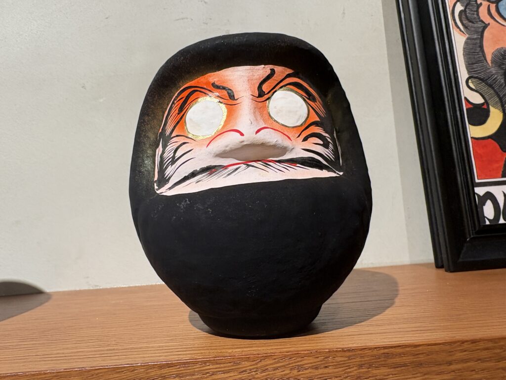 A large black daruma doll with traditional orange facial features and blank white eyes displayed on a wooden shelf.