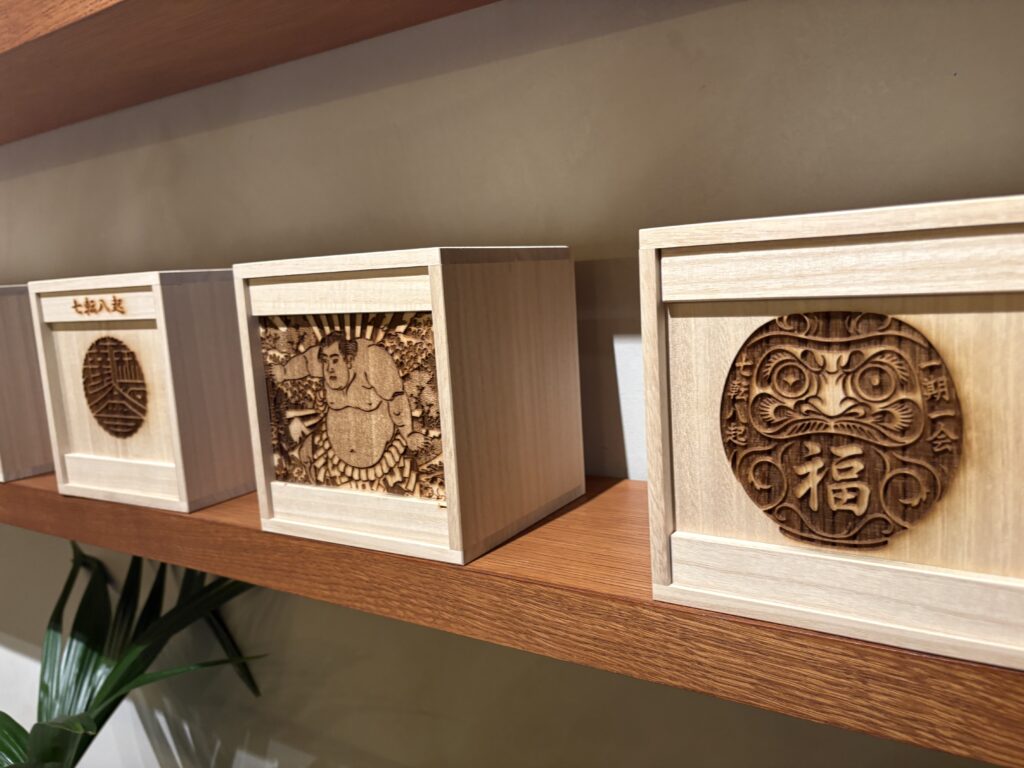 Wooden display boxes with intricate carved designs including daruma faces and traditional Japanese motifs arranged neatly on a shelf.