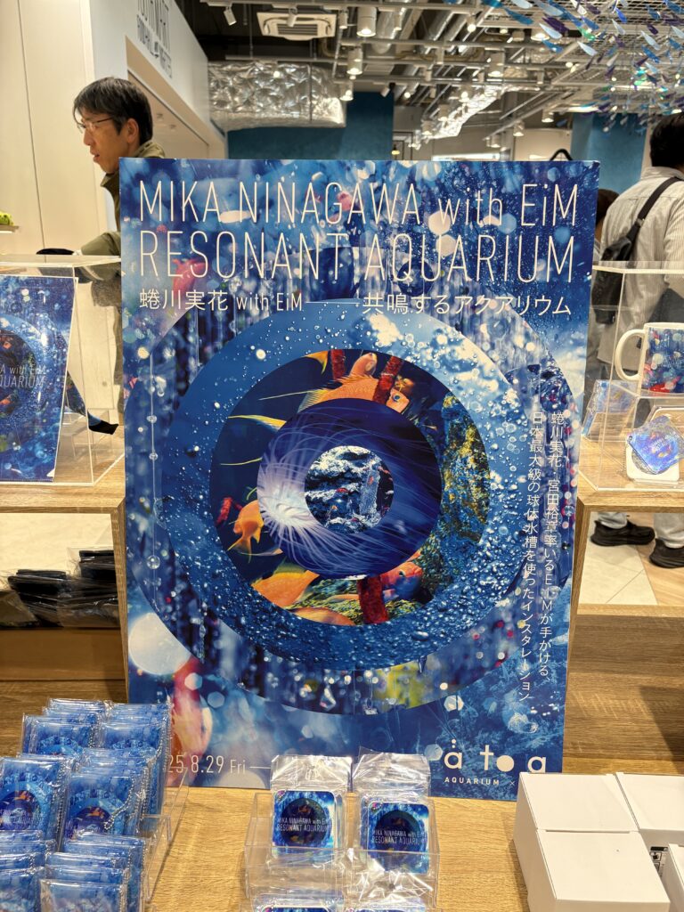 A vibrant promotional poster for Mika Ninagawa with EiM “Resonant Aquarium,” displayed inside a gift shop surrounded by blue-themed merchandise.