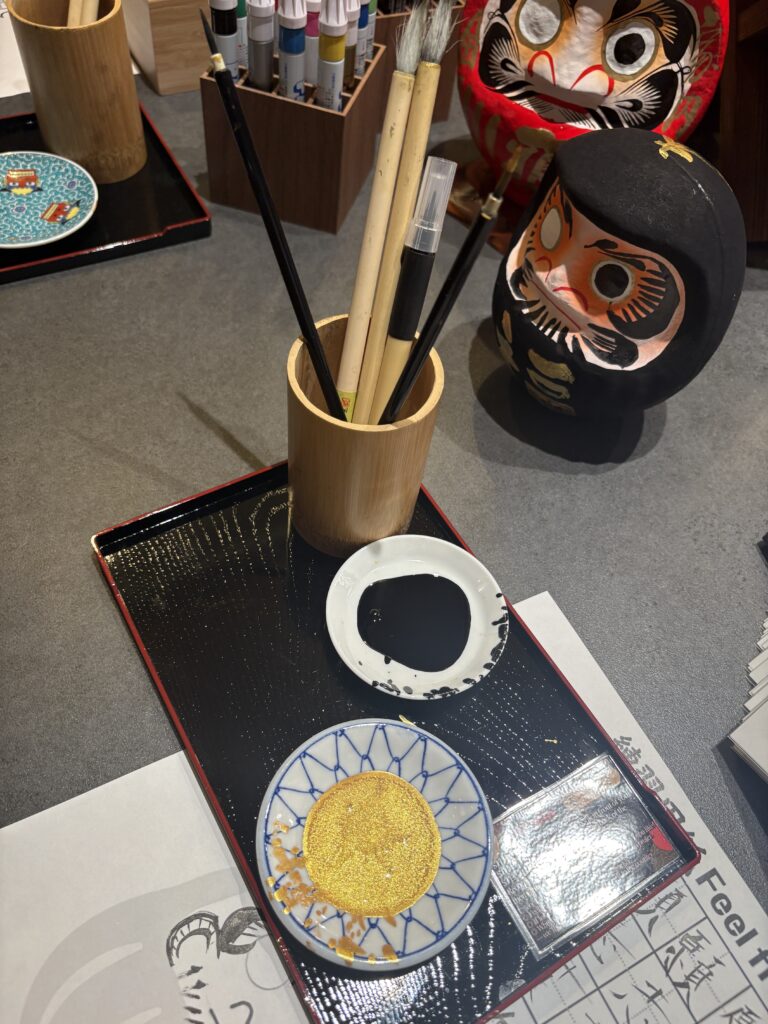 Painting tools including brushes, ink, gold powder, and small plates arranged on a tray beside a partially painted black daruma doll.