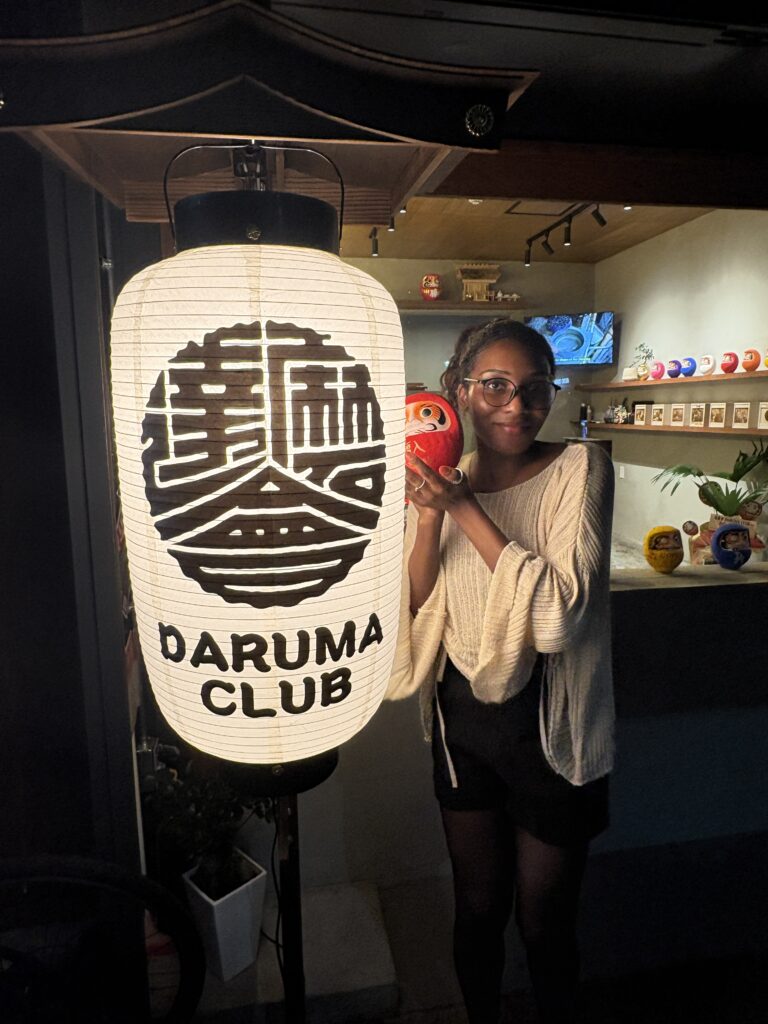 A person standing outside Daruma Club at night, holding a red daruma doll beside a large illuminated lantern with the Daruma Club logo.
