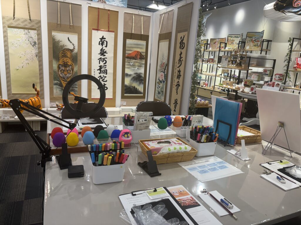 A workshop table with colorful blank daruma forms, markers, tools, and Japanese hanging scrolls in the background.