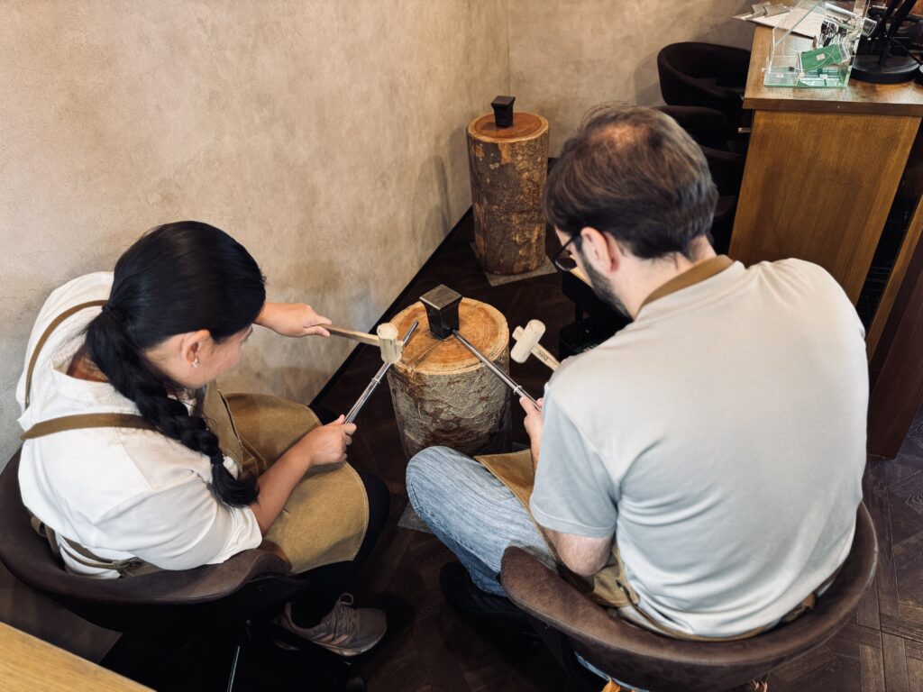 Two people hammering metal strips on a wooden stump during the ring crafting process at Début icci Daikanyama.