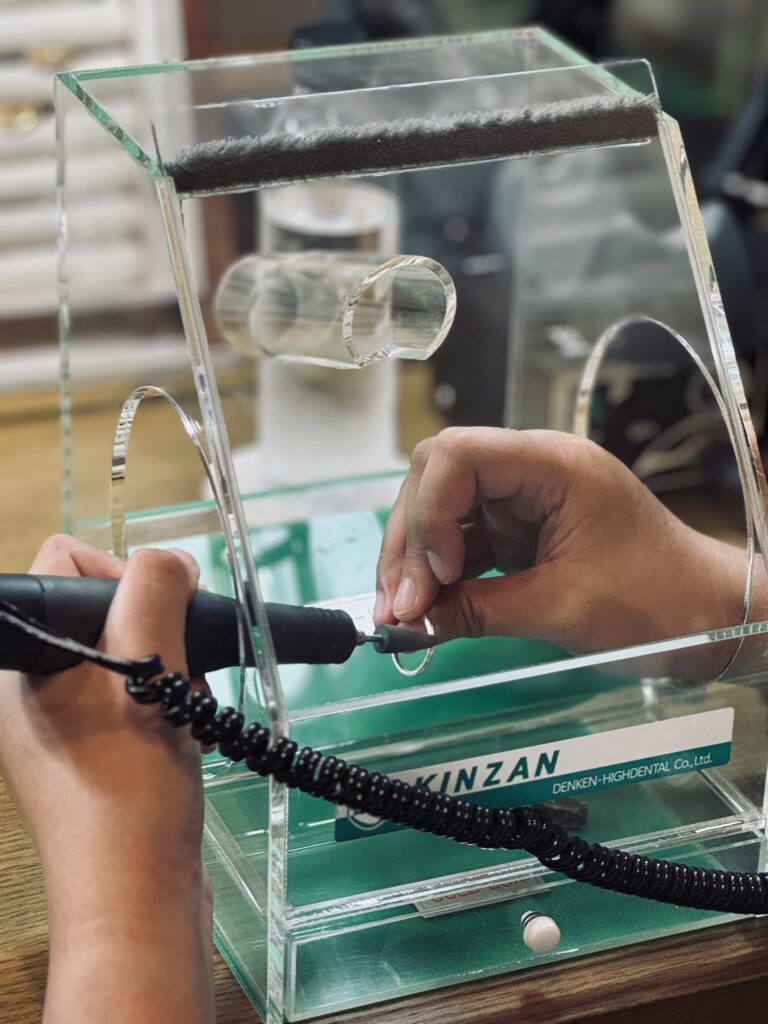 A close-up of hands polishing a ring using an electric tool inside a transparent protective box.