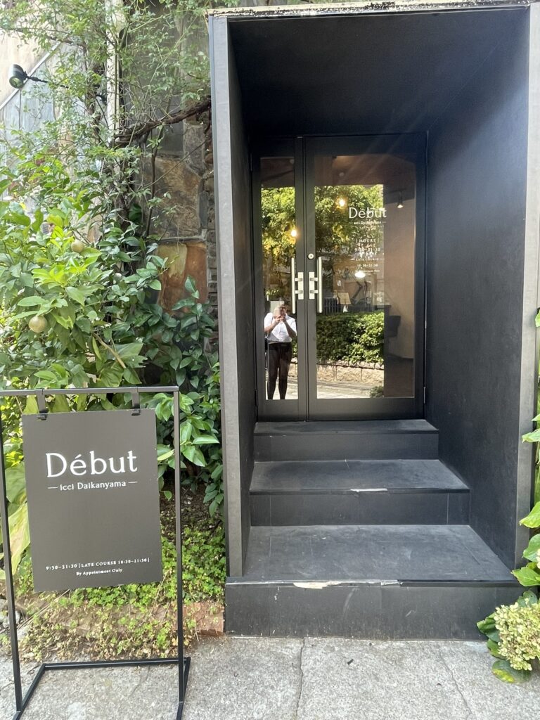 The entrance of Début icci Daikanyama with a black doorway and a sign displayed outside surrounded by greenery.