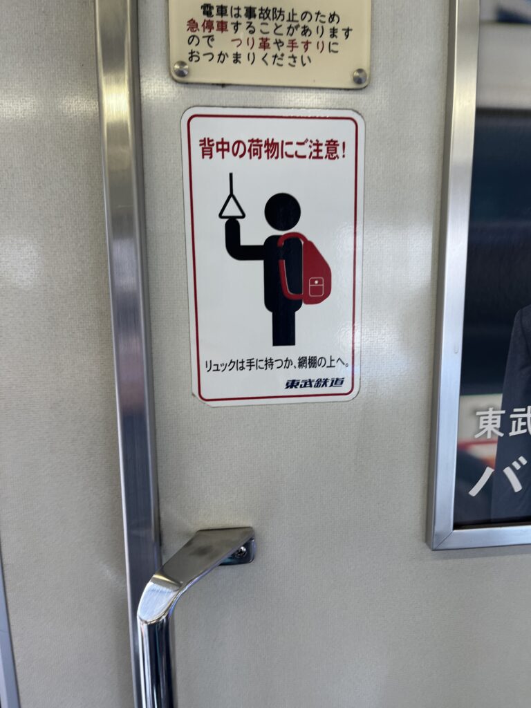 A Japanese train sign reminding passengers to be careful with backpacks, showing a person holding a strap with a red backpack worn on the back.