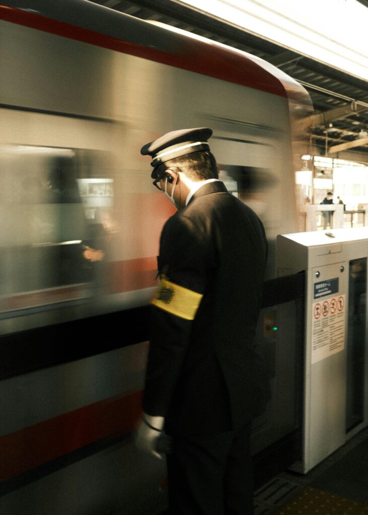 A Japanese station attendant in uniform standing beside a moving train, ensuring safety as it departs.