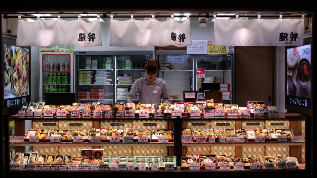 A shopkeeper arranging colorful bento boxes and onigiri at a Japanese ekiben store.