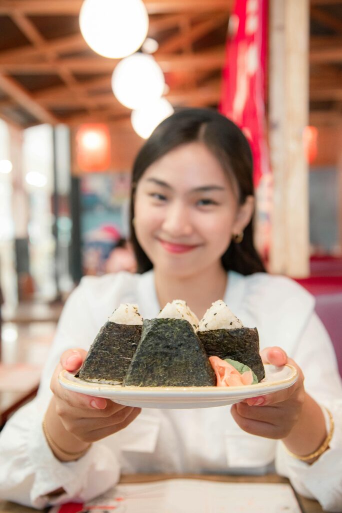 A smiling woman holding a plate of three freshly made onigiri in a Japanese restaurant.