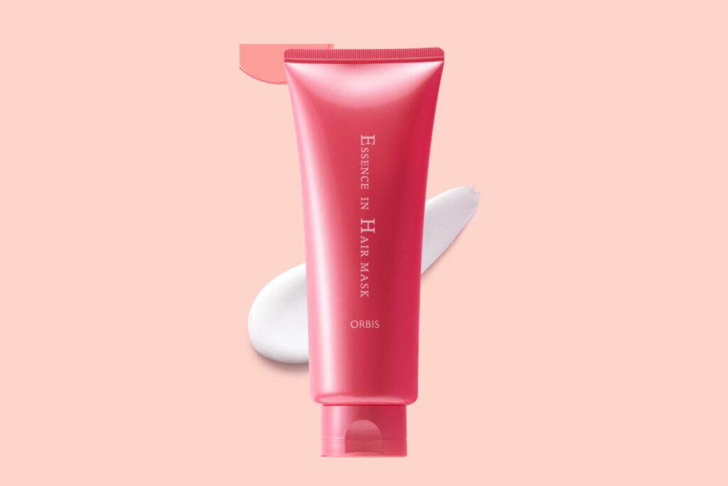 Orbis Essence in Hair Mask in a pink tube displayed on a soft peach background with creamy texture beside it.