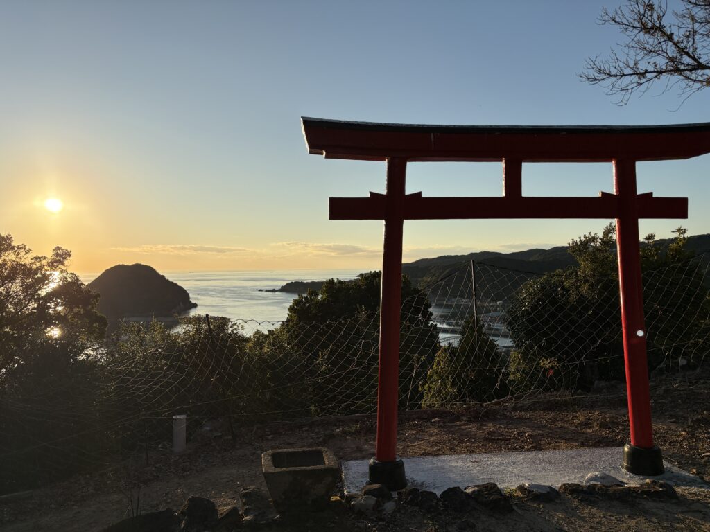 A red torii gate overlooking the ocean at sunset in Susami, Wakayama, with warm golden light reflecting on the water and mountains in the distance.