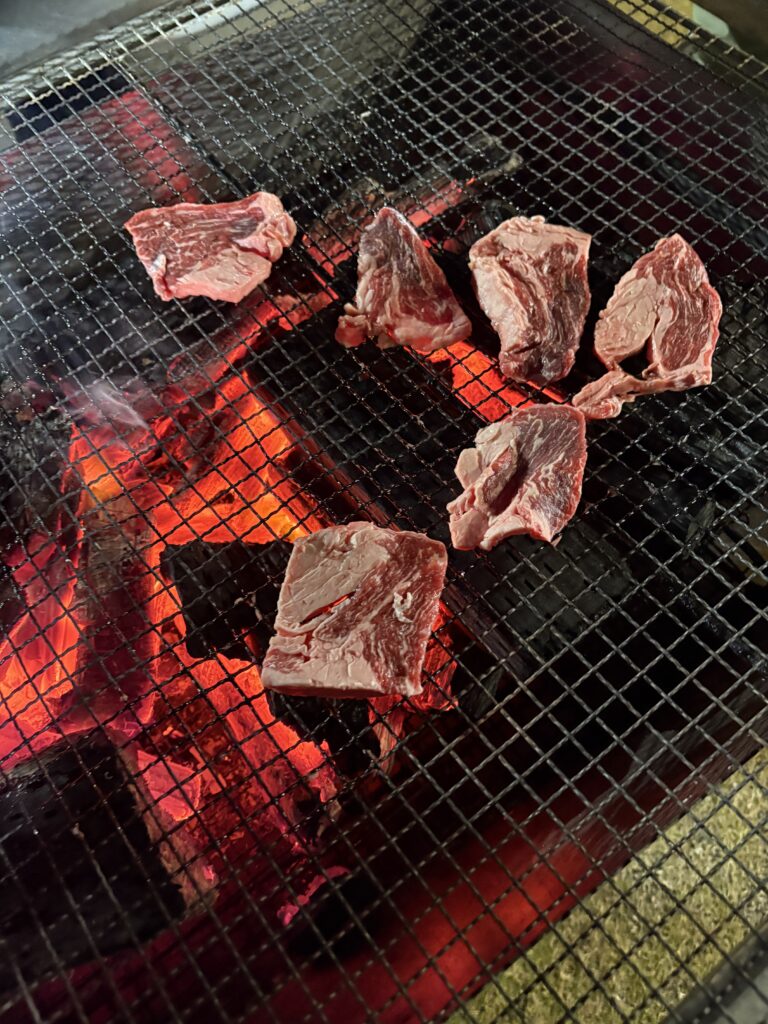 Slices of raw meat cooking over glowing charcoal on a metal grill during a beachside yakiniku dinner in Susami.