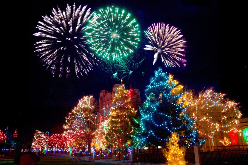 A vibrant night scene featuring colorful Christmas trees covered in lights, with large fireworks bursting in the sky above a historic building.
