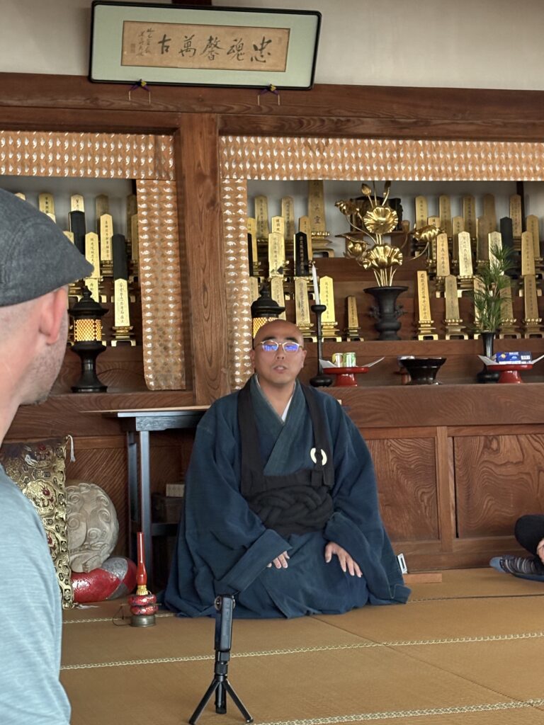 A Buddhist monk in traditional robes teaching zazen meditation inside a wooden temple decorated with gold ornaments and ancestral tablets.