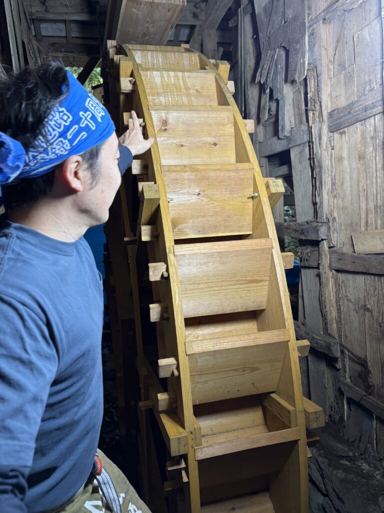 A man wearing a blue bandana explaining a traditional wooden incense watermill inside an old wooden structure in Susami.
