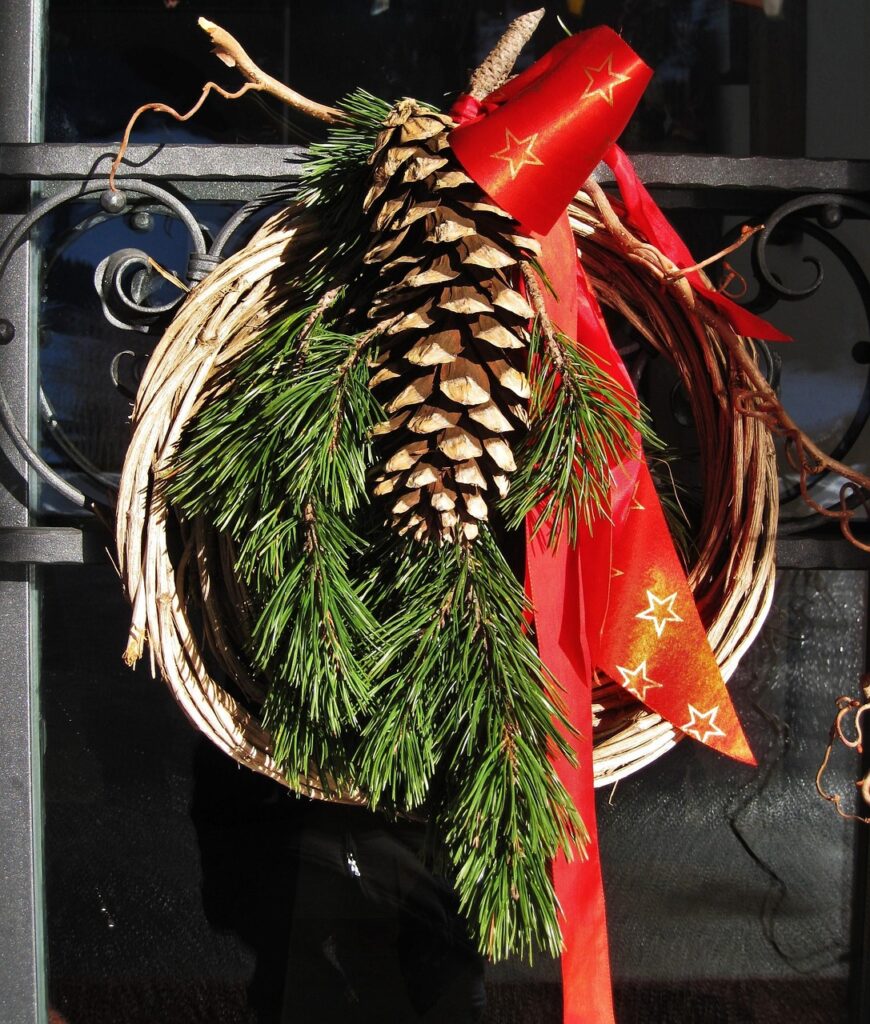 A rustic Christmas wreath decorated with pine branches, a large pinecone, and a red ribbon with gold stars.