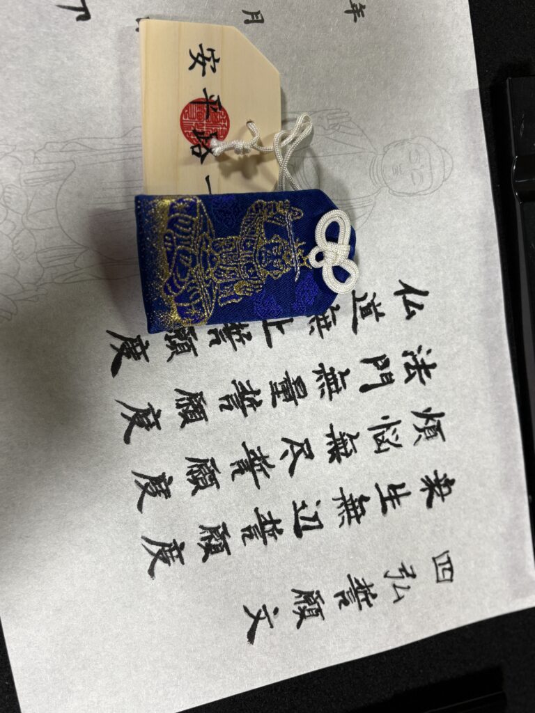 A close-up of a completed sutra copying sheet with black ink characters, alongside a blue omamori charm and wooden prayer plaque.