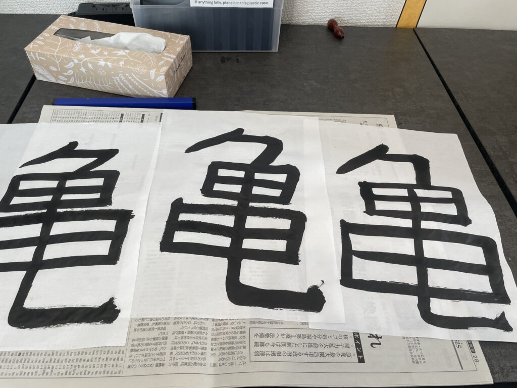 Sheets of paper displaying large black kanji characters laid out on a table during a Japanese calligraphy workshop.
