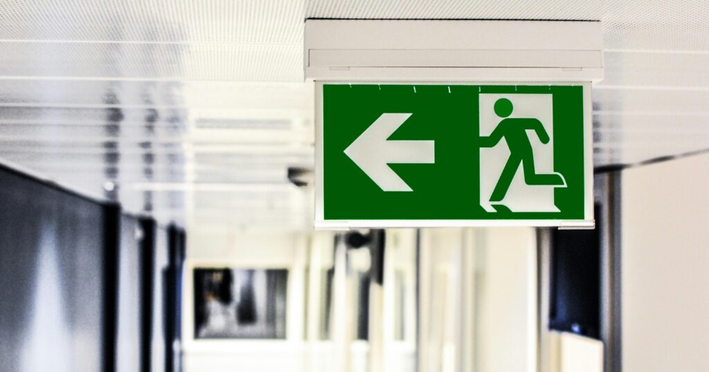 Green emergency exit sign with a running figure and an arrow pointing left in a bright hallway.