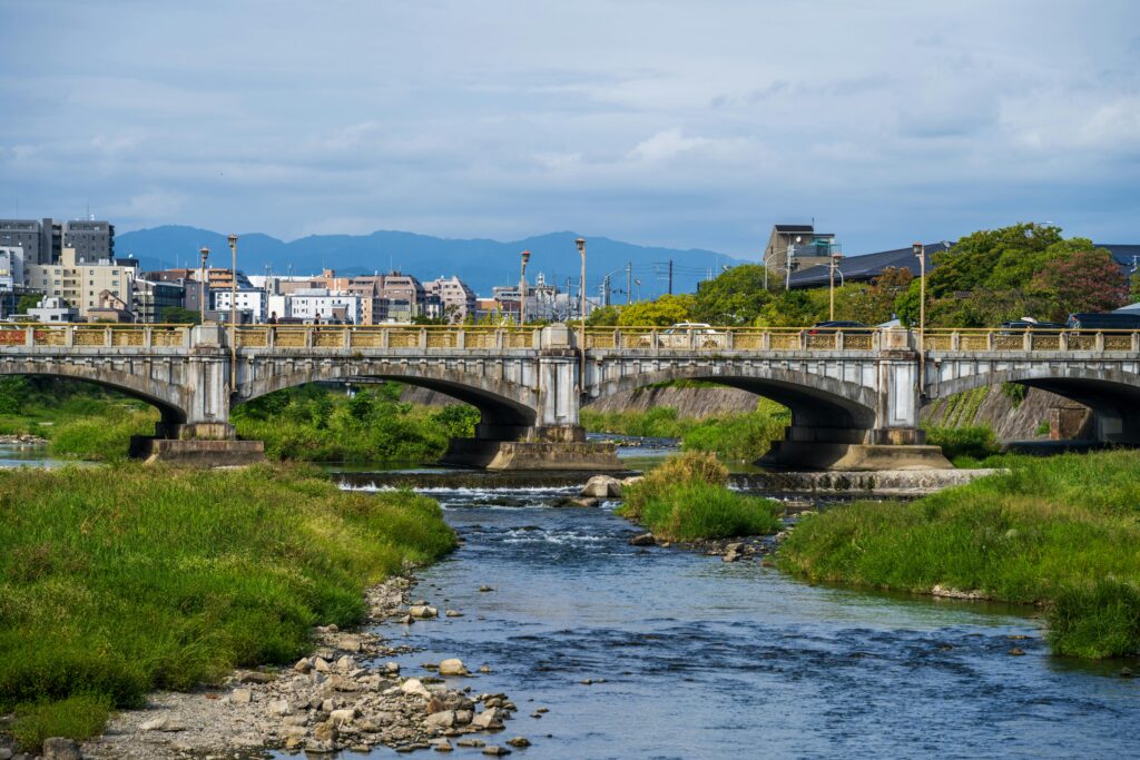 Calm river flowing under an old concrete bridge with buildings and mountains in the background.