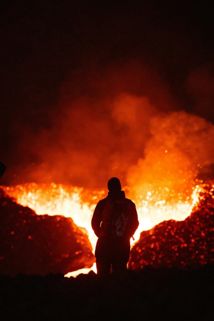 Silhouette of a person standing near the glowing lava of an erupting volcano at night.