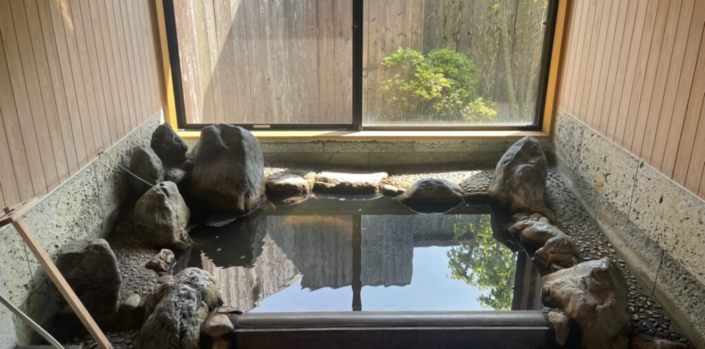 A private indoor onsen bath surrounded by natural stones with calm water reflecting the window light.