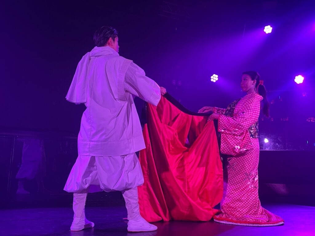 Performers in traditional Japanese costumes reenact a dramatic scene on stage under purple lighting during a cultural performance.
