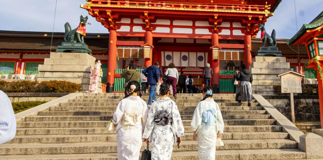 Three women wearing pastel-colored kimonos walk up the stone steps toward a bright red Shinto shrine gate in Japan.