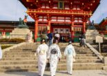 Three women wearing pastel-colored kimonos walk up the stone steps toward a bright red Shinto shrine gate in Japan.