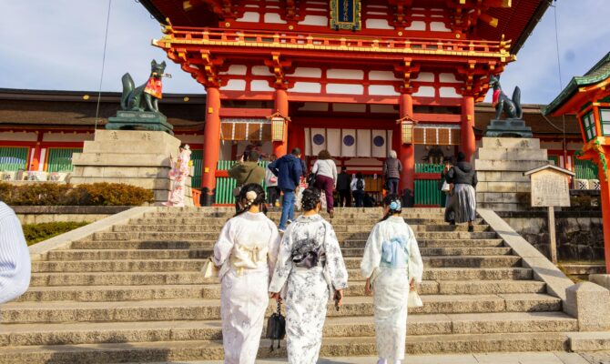 Three women wearing pastel-colored kimonos walk up the stone steps toward a bright red Shinto shrine gate in Japan.