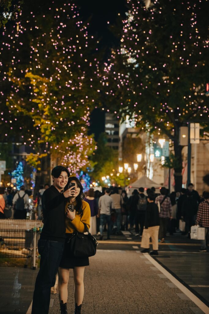 Couple taking a selfie on a busy Japanese street decorated with festive lights during the New Year season at night.