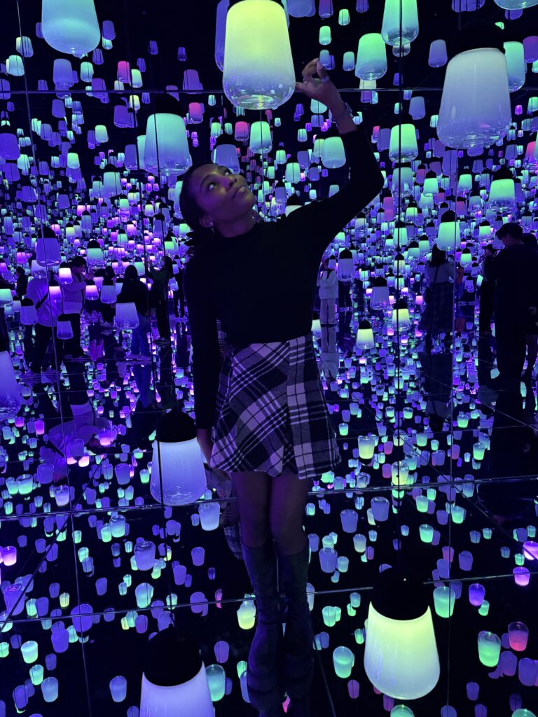 A visitor standing inside an immersive room filled with warm amber lanterns reflected endlessly by mirrored walls and floors.