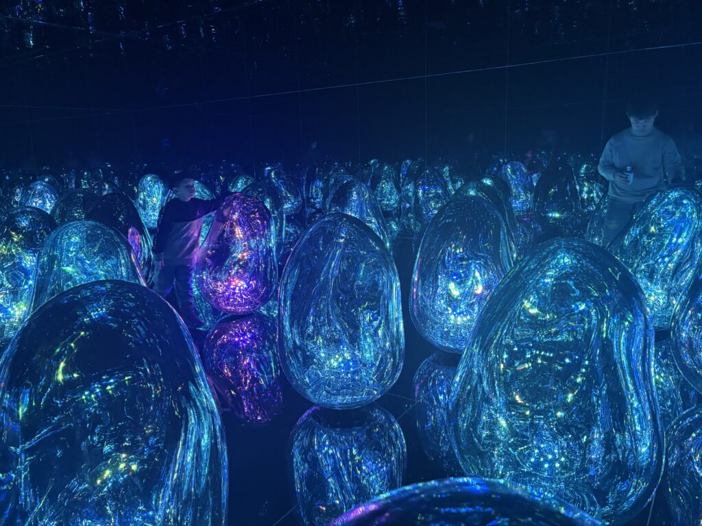 Visitors walking among large, glowing crystal-like light sculptures in shades of blue and purple, reflected endlessly by mirrored floors and walls inside a teamLab installation.