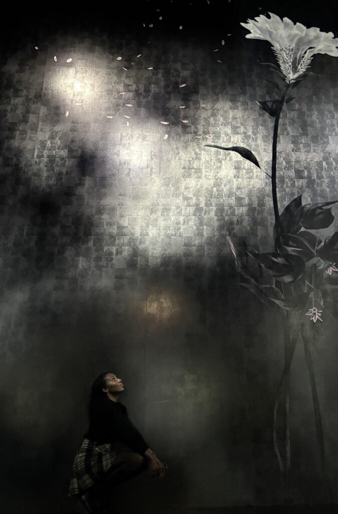 A large digital floral artwork showing a tall white flower and drifting petals against a dark, textured background, with a visitor seated below looking upward.