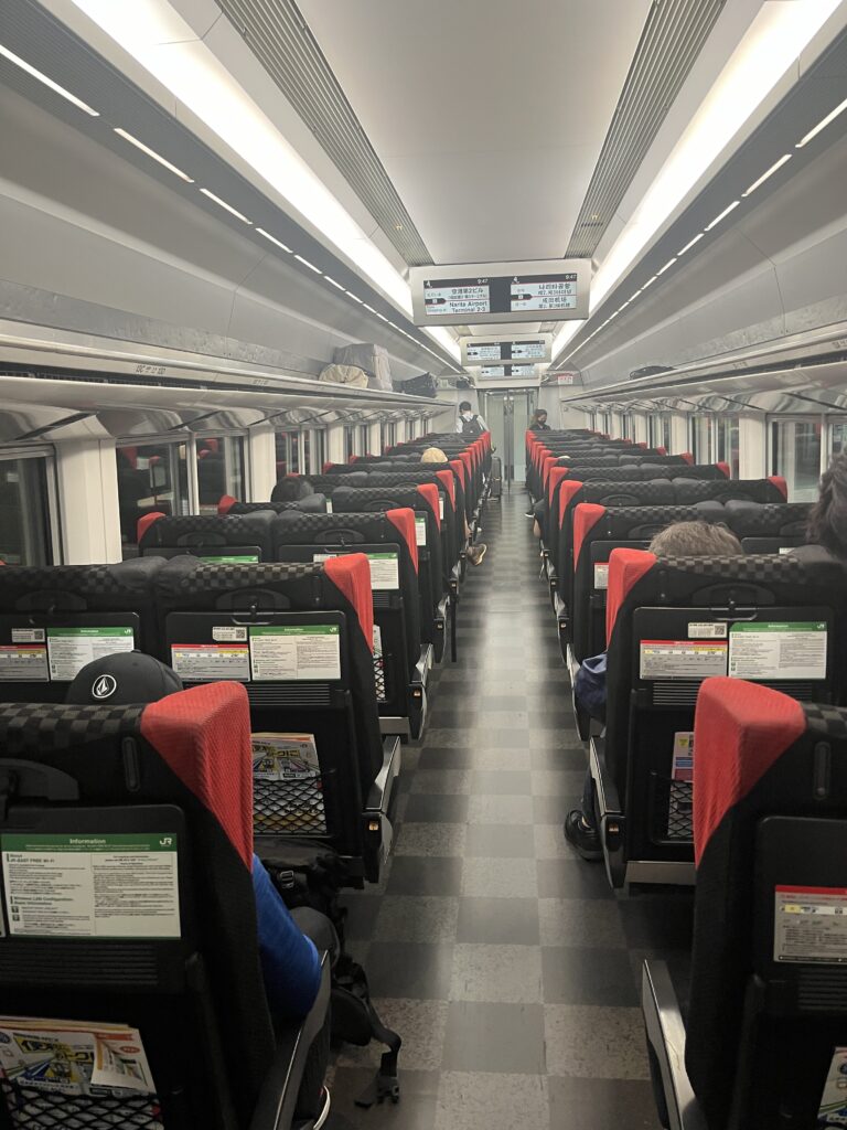 Interior of the Narita Express train showing rows of black and red seats, overhead luggage racks, and passengers seated during the ride to or from the airport.