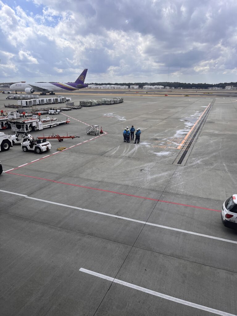 View of Narita Airport’s tarmac with ground crew standing near cargo containers and service vehicles, with a Thai Airways aircraft parked in the background.