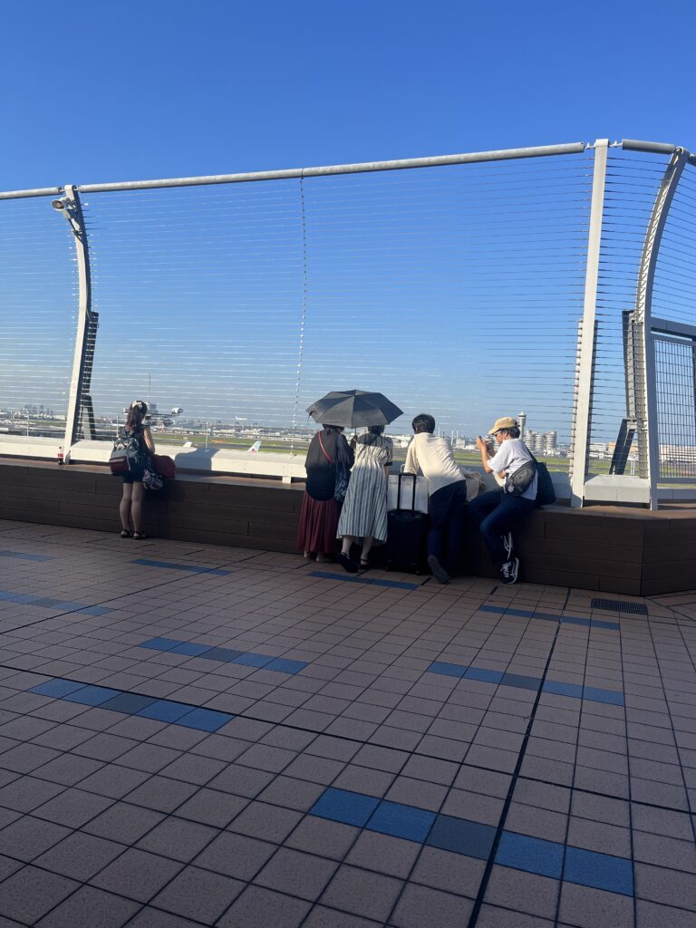 Observation deck at Haneda Airport with people watching airplanes take off and land behind a safety fence under a clear blue sky.