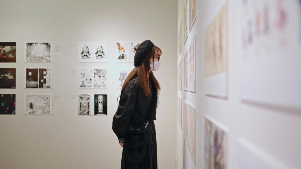 Visitor wearing a mask and beret closely examining framed illustrations in a quiet contemporary art gallery.
