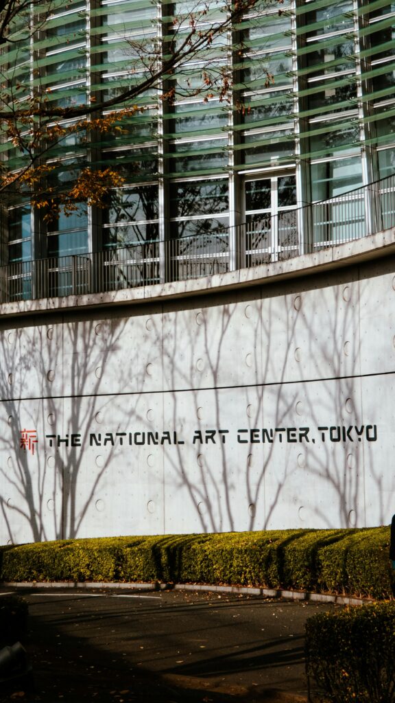 Exterior wall of the National Art Center, Tokyo, with modern glass architecture and tree shadows cast across the facade.