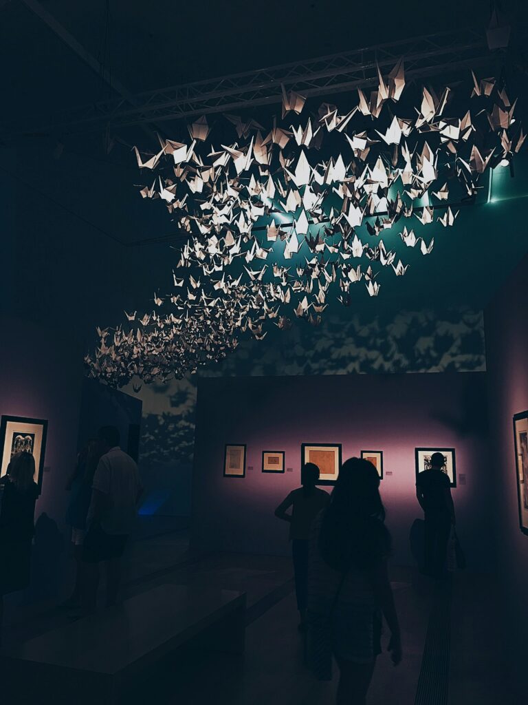 Art installation of hundreds of suspended origami paper cranes floating above visitors inside a dimly lit exhibition space.
