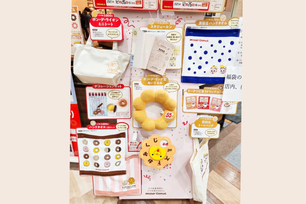 Mister Donut fukubukuro display showing tote bags, donut-themed towels, planners, plush items, and drink vouchers arranged on a store board.
