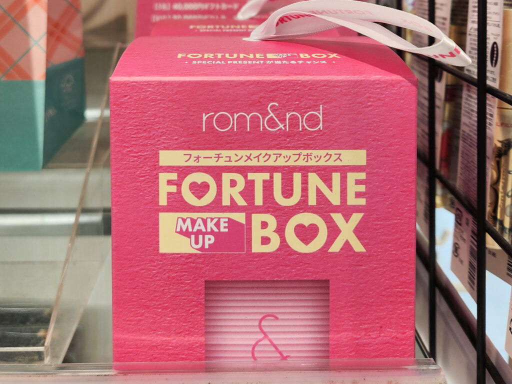 Pink rom&nd Fortune Makeup Box fukubukuro displayed on a cosmetics shelf, labeled as a mystery makeup lucky box.