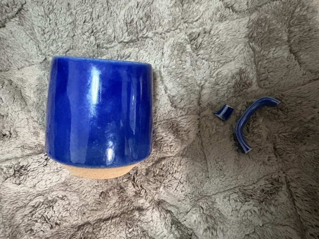 Broken blue ceramic mug with detached handle pieces laid out on a soft surface before repair.